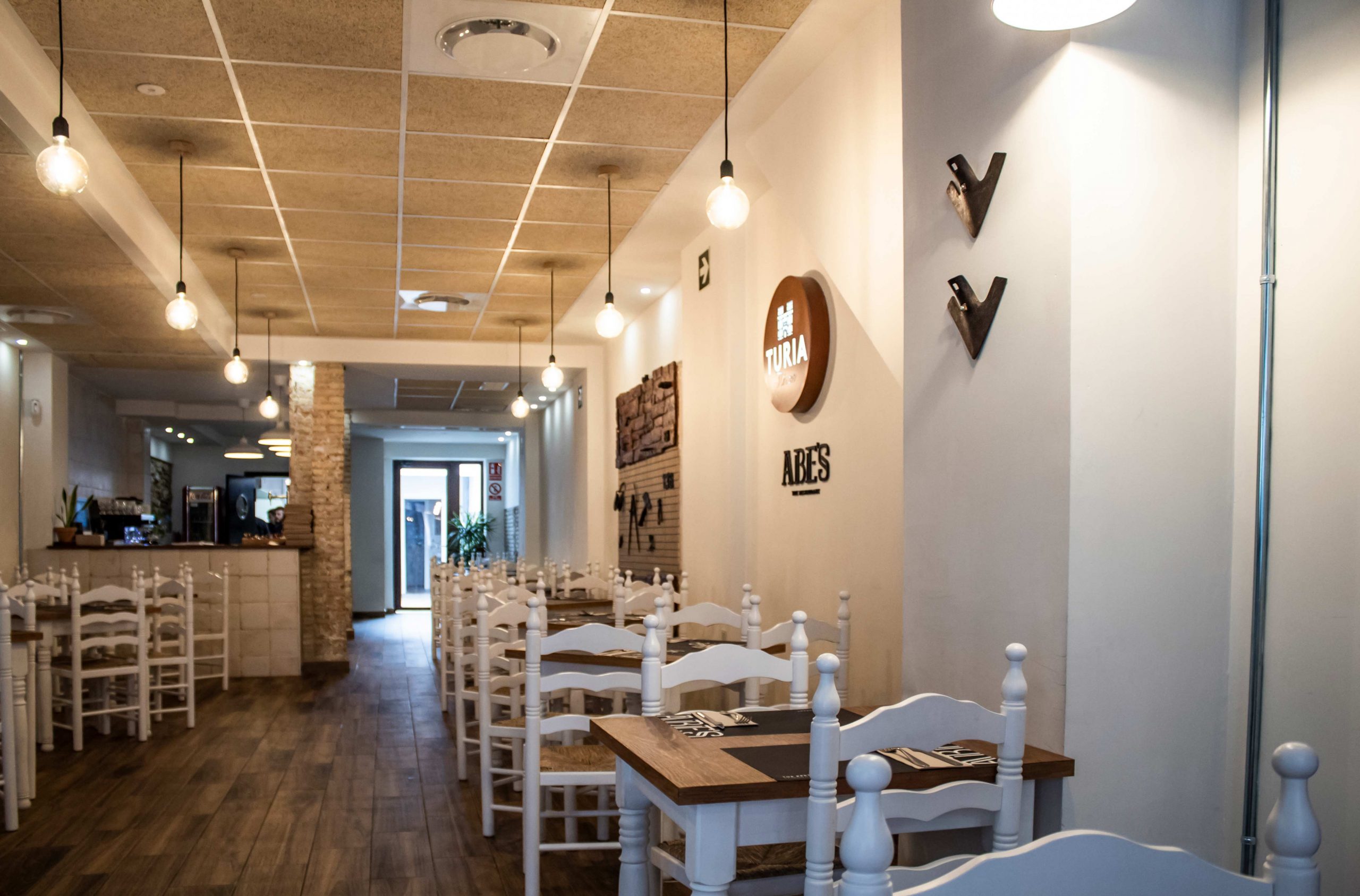 Interior de Abe's en Aldaia, con mesas de madera, sillas blancas y el logo en la pared